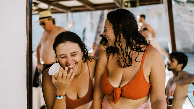 Two women in swimsuits laughing on a boat with people in the background.