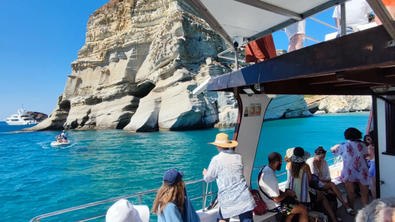 People on a boat near a rocky coastal cliff with turquoise water and a yacht in the background.
