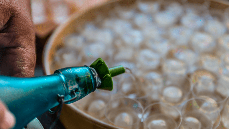 Hand pouring liquid from blue bottle into small plastic cups on a tray.