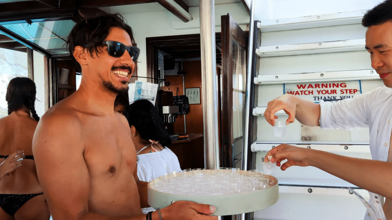 Man holds tray of drinks, others take drinks on a boat near stairs.