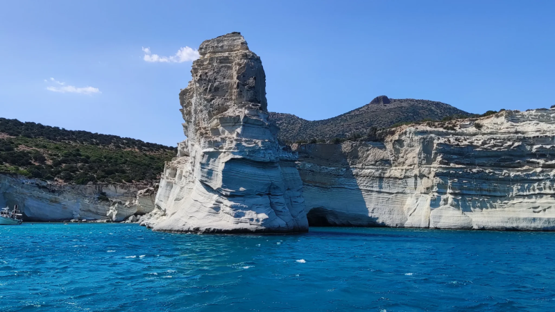 Tall, rocky sea cliff with clear blue waters and a boat in the distance under a clear blue sky.