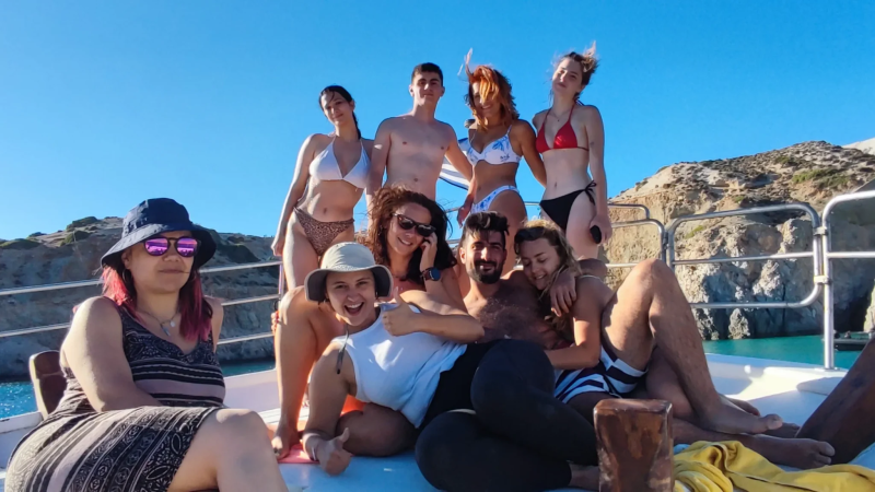 Group of people in swimsuits posing on a boat with rocky coast background.