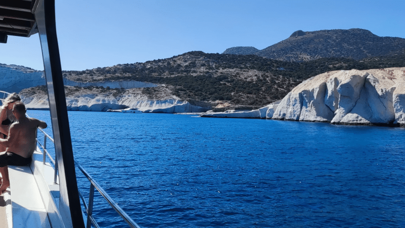 People on a boat near rocky coastline with blue sea and clear sky.