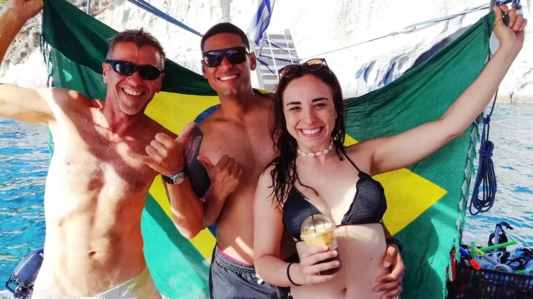 Three smiling people on a boat holding a Brazilian flag with a seaside backdrop.