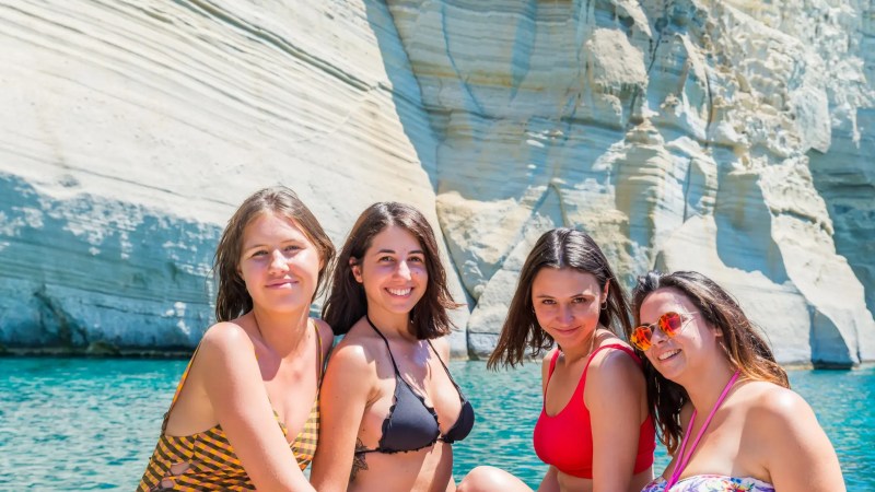 Four women in swimsuits smiling by a rocky cliff at a seaside location.