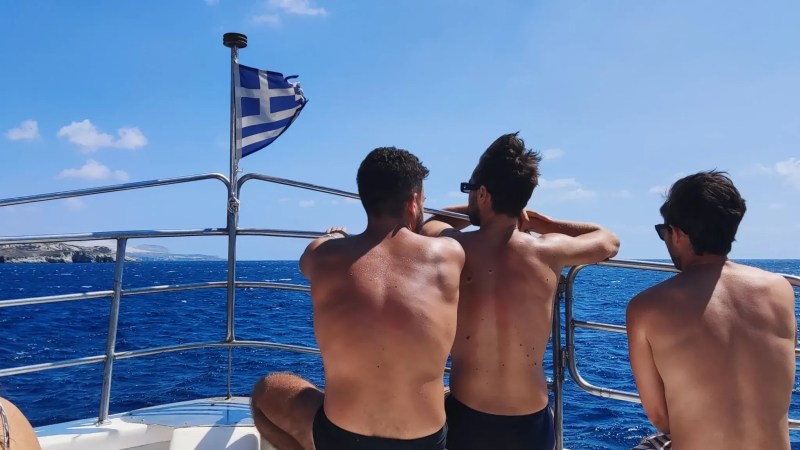 Three people on a boat with Greek flag, enjoying sunny sea view.
