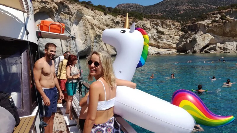 People on a boat with a large unicorn float, near cliffs and swimmers in blue water.